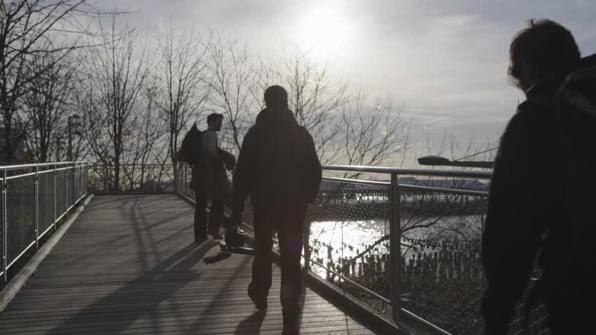 Male friends walk over a fenced footbridge carrying equipment on an overcast winter day