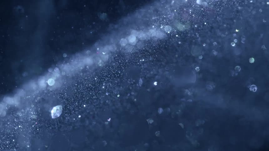 Glittery particles swirl around in a shimmering almost weightless motion reflecting bright flecks of light across a dark backdrop while countless specks dance and sparkle in a mesmerizing luminous pattern