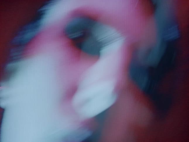 A grainy stylized shot depicts a woman with short dark hair moving her head in front of a deep red background featuring a double exposure