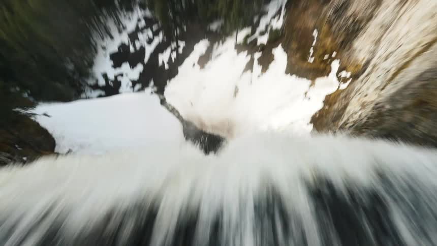 A waterfall plunges down a rocky cliff face partially covered in snow as seen from an aerial perspective