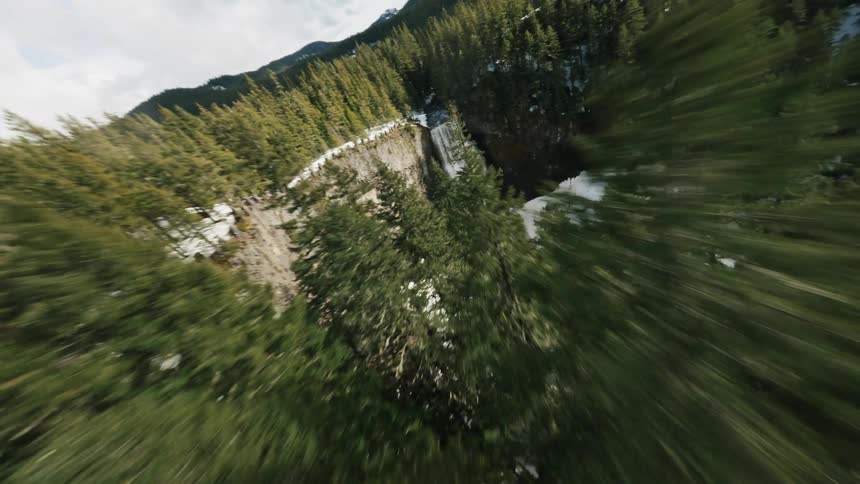 A dense forest of evergreen trees on a steep hillside partially obscuring a waterfall cascading down a rocky cliff face in the background