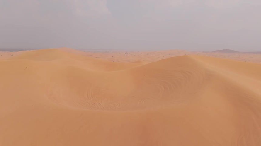 From an elevated vantage vast stretches of orange sand dunes extend across the frame with subtle ridges and scattered patches of sparse vegetation leading toward a hazy horizon under a muted sky