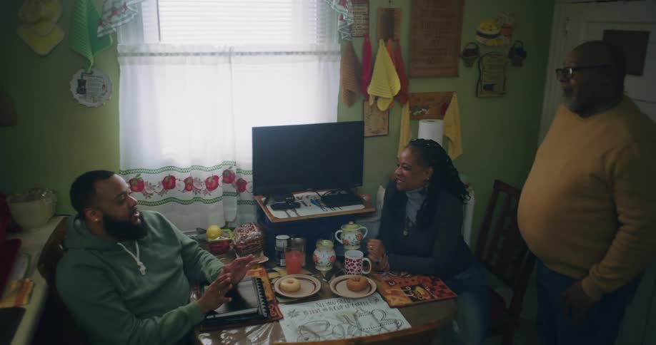 A man in a green hoodie talks at a table in a kitchen with two other people while a tv sits on a small shelf
