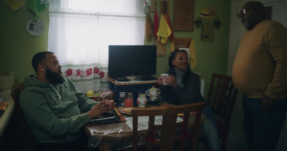 A couple sits at a table in a home kitchen as they talk to a standing man