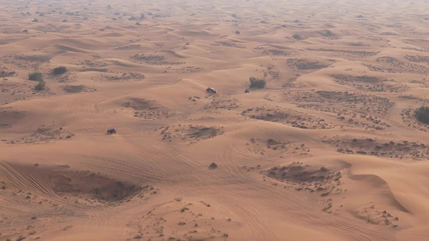 An aerial view shows vehicles driving on a vast undulating landscape of sandy dunes with sparse vegetation under a hazy sky