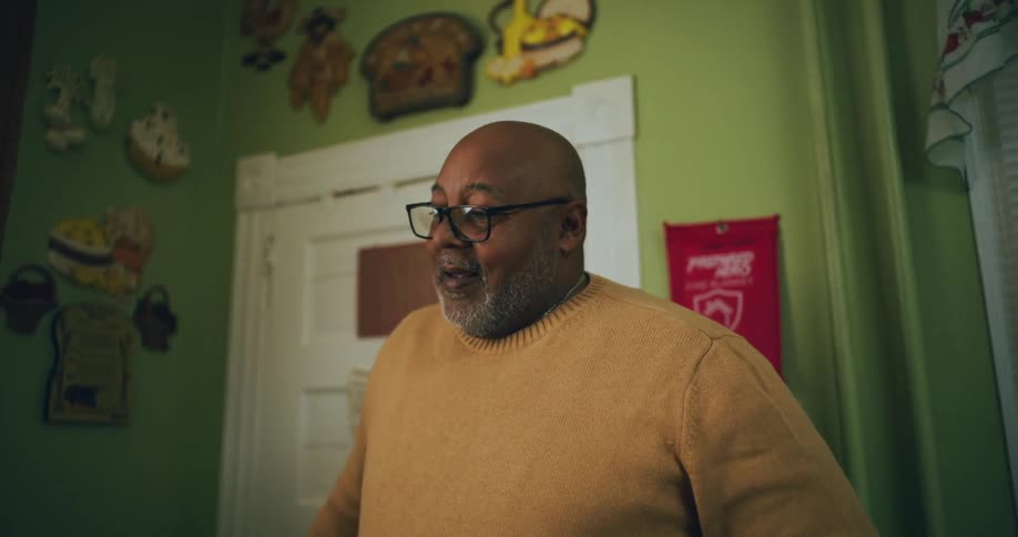 A man wearing a yellow sweater standing on a room at home talking to someone