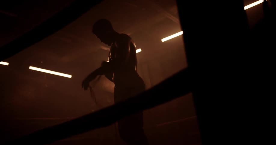 Boxer standing in a boxing ring in a dimly lit gym unwinds the wrapping on his hands