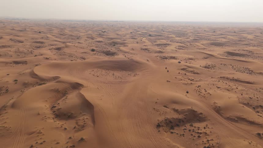 Multiple offroad vehicles traverse sweeping desert dunes from a high vantage with ridges stretching across the horizon faint tire tracks marking the sand and scattered shrubs visible under a hazy sky