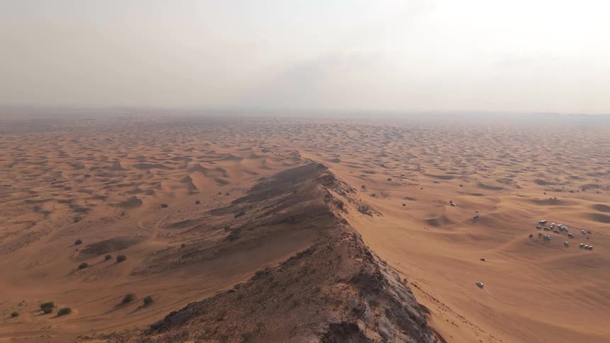 Aerial view over vehicles driving through the valley of a large sand dune at the edge of a vast desert under a foggy clouded sky