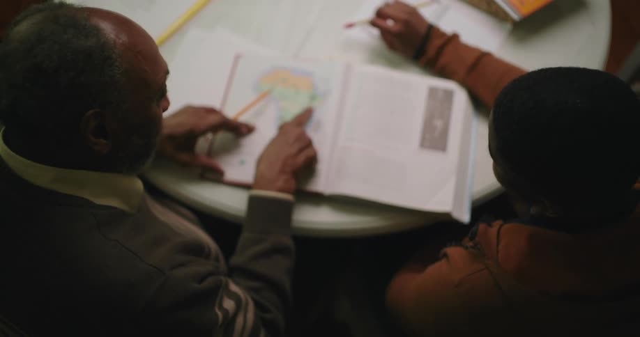 An adult points to a map in a book while assisting a young boy with his homework