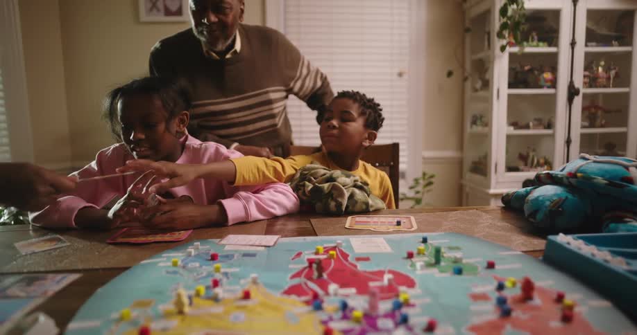 A family is gathered around a table playing a boardgame with a colorful map at home