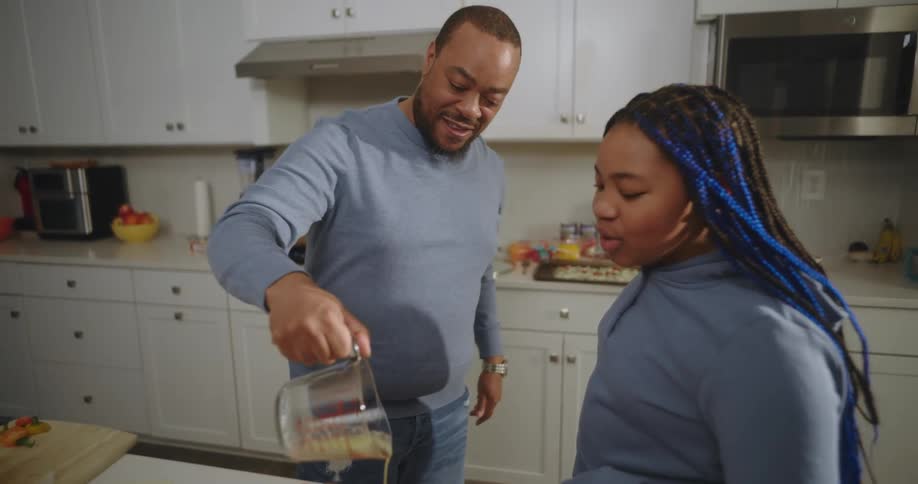 A man is preparing food in a home kitchen while a woman watches and talks to him