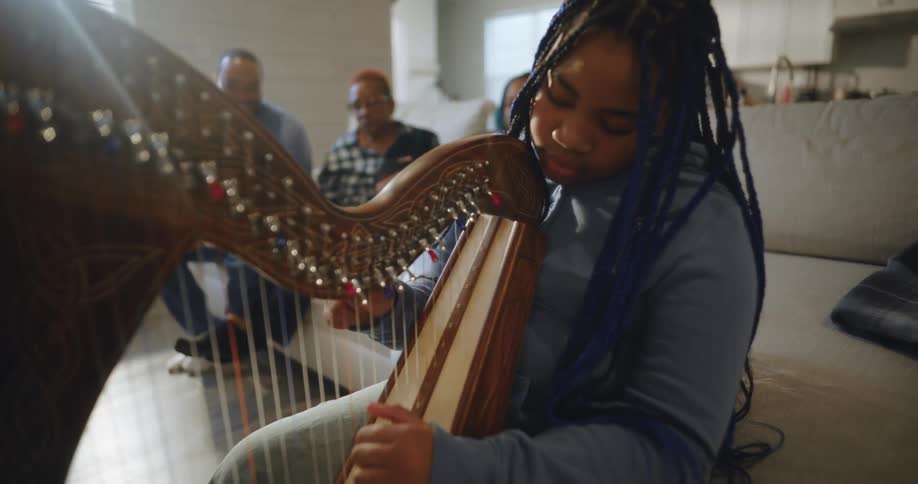 A teen plays a harp in the foreground while her family sit on a couch in the background
