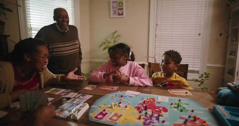 A family is gathered around a table playing a board game with a colorful map and various game pieces while some are holding cards and discussing their moves
