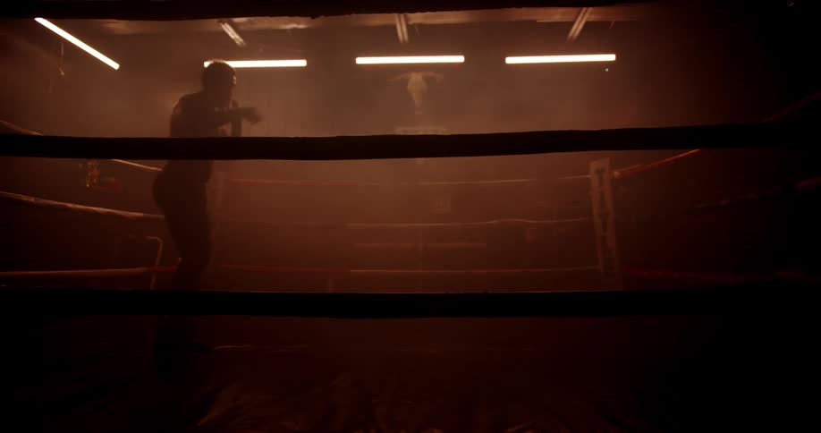 A boxer trains alone in a dimly lit gym practicing on the ring