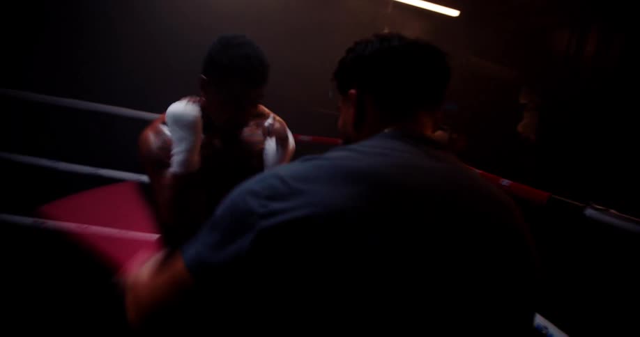 A boxer trains with a sparring coach in a dimly lit gym practicing on the ring
