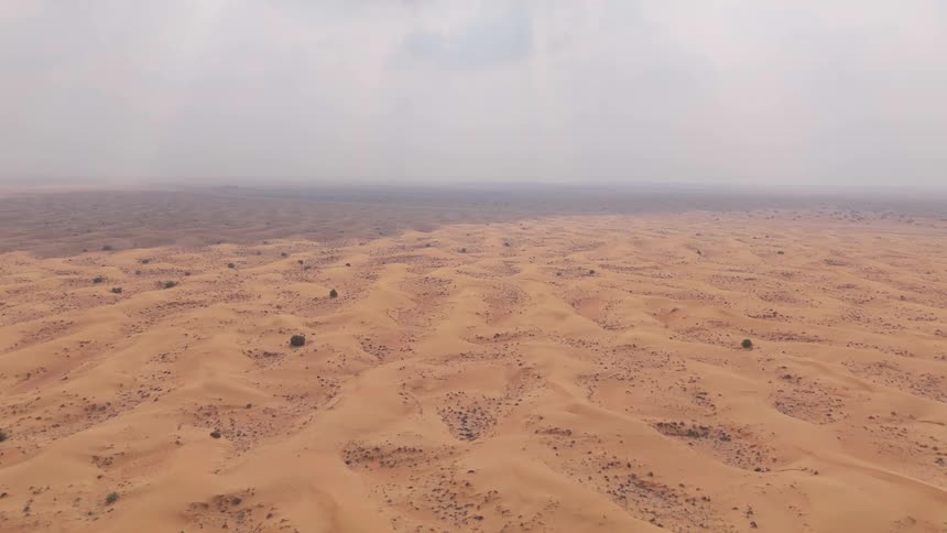From a steadily rising vantage vast orange dunes and sparse vegetation stretch across the horizon under a hazy sky with shifting shadows cast by passing clouds over the rolling sands