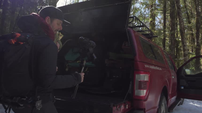 A father carries a child and camera equipment on a stand near a red pickup truck in a forested area surrounded by tall trees and scattered foliage