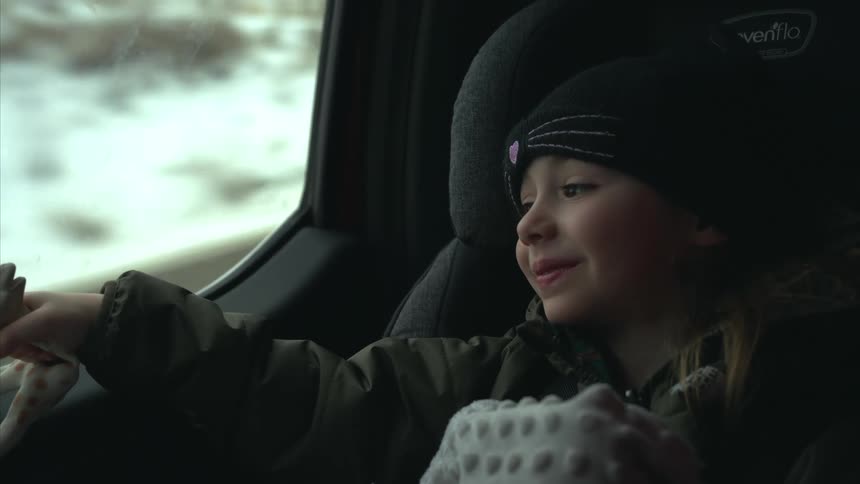 Cheerful little girl sitting in a baby seat playing with a toy while in a moving car