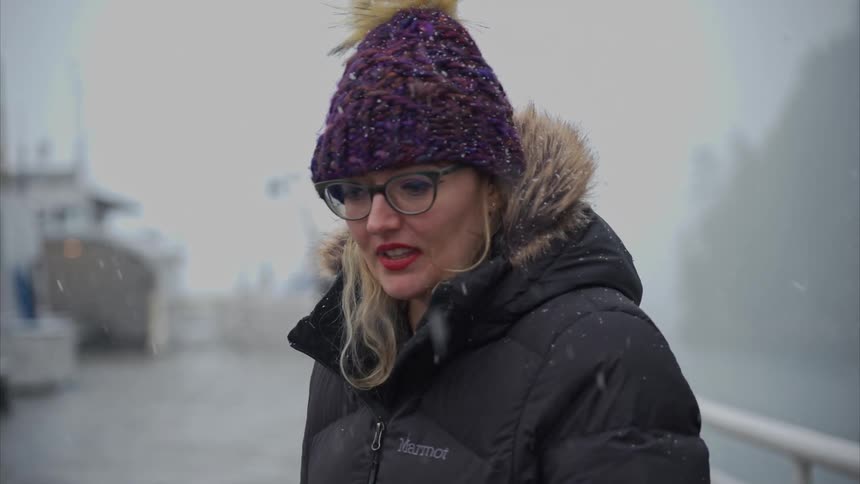 A woman in a purple knit hat with a yellow pompom and a black jacket stands on the snowy deck of a ship with a misty forest in the background
