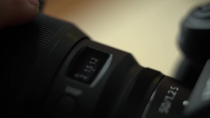 In a closeup view a persons hand carefully rotates the focusing ring of a camera lens to finetune the fstop while a small digital display updates in real time and the softly blurred background emphasizes the subtle adjustments