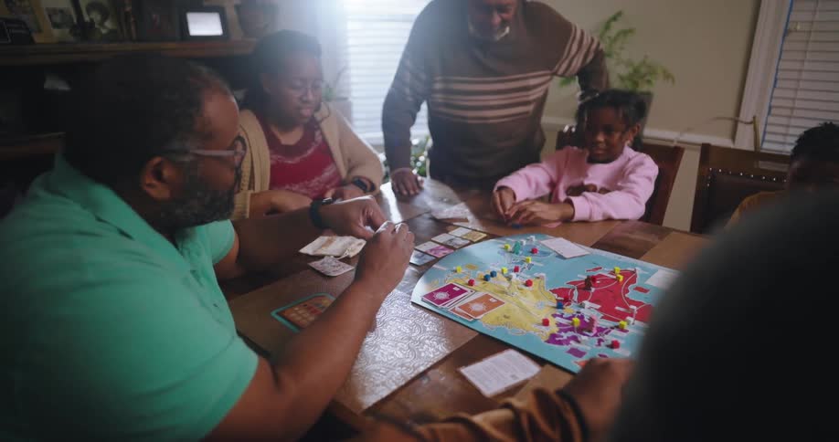 The family is sitting around the dinner rooming table playing a board game while the grandfather is helping the young girl play