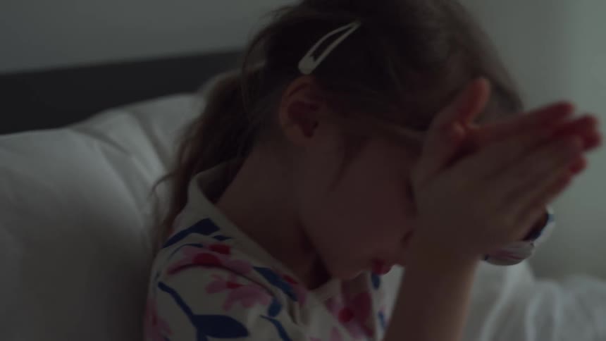 A girl sits on a bed in a dimly lit bedroom wearing pajamas and gesturing as she talks