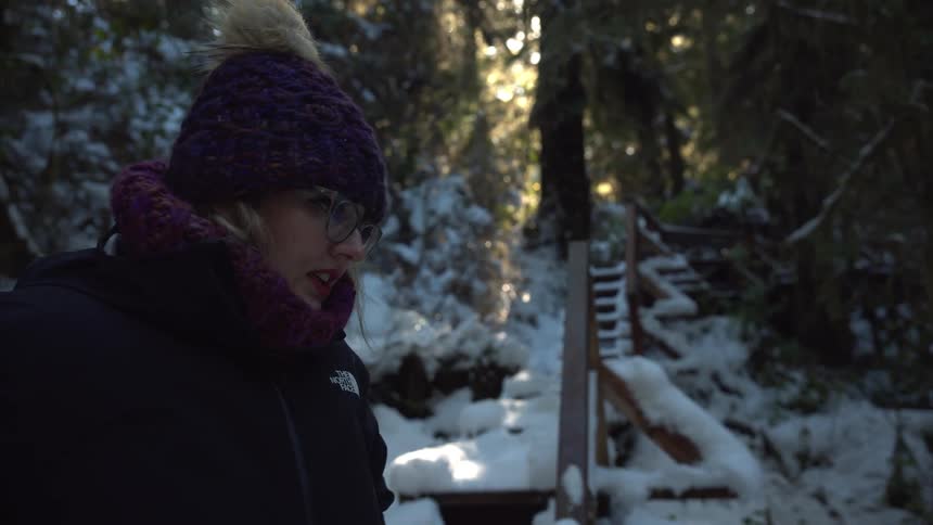 A woman wearing a purple knit hat with a pompom and a winter jacket stands in a snowy forest with a wooden staircase in the background