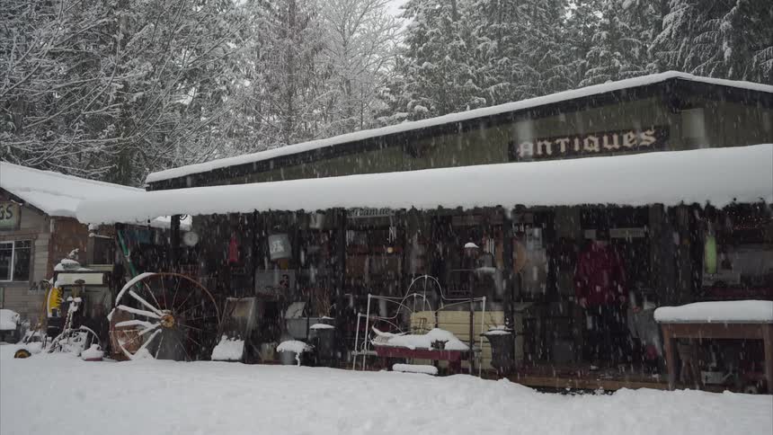 A child runs out of a rustic antiques store followed by her father as snow falls gently around them with a pine tree forest in the background
