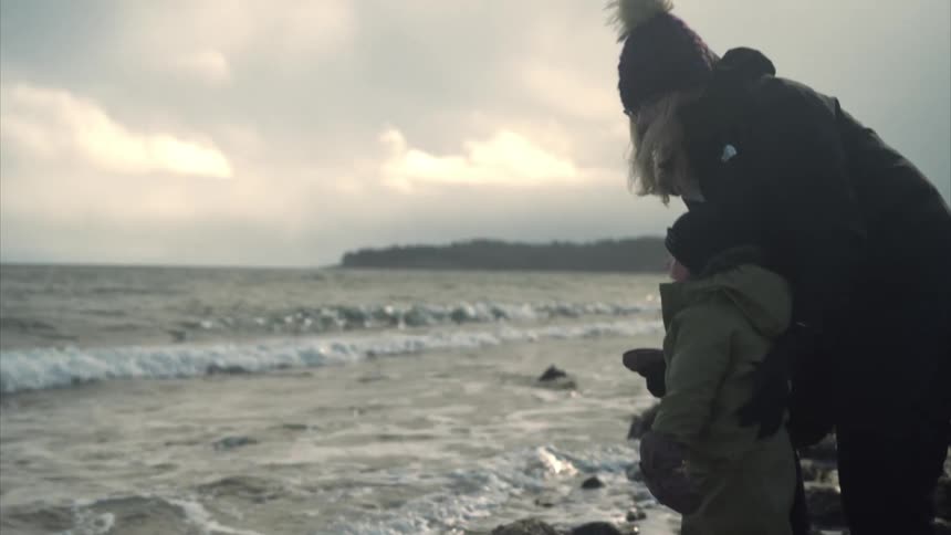 Daughter dressed in a warm snowsuit and gloves stands on a rocky shoreline reaching out toward the waves as mother gently holds them for support
