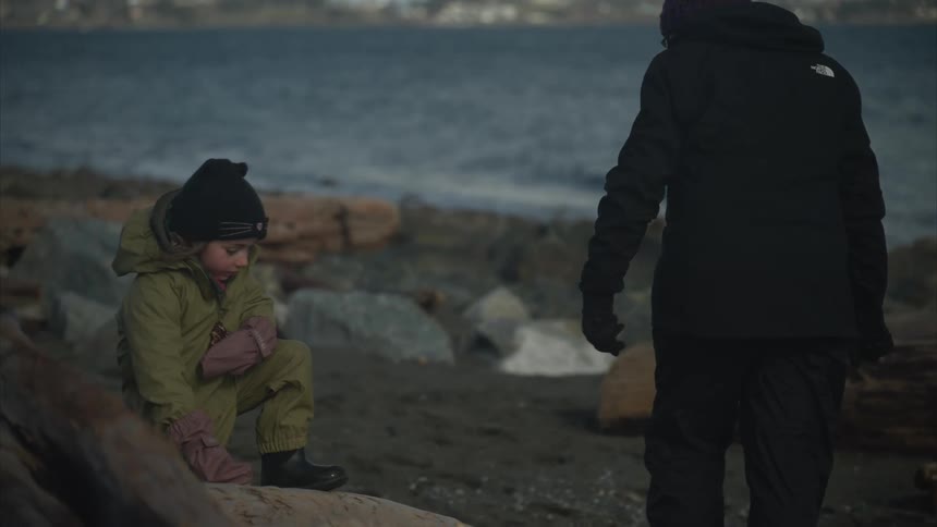 A mother and daughter bundled in thick winter clothes walk along a rocky beach