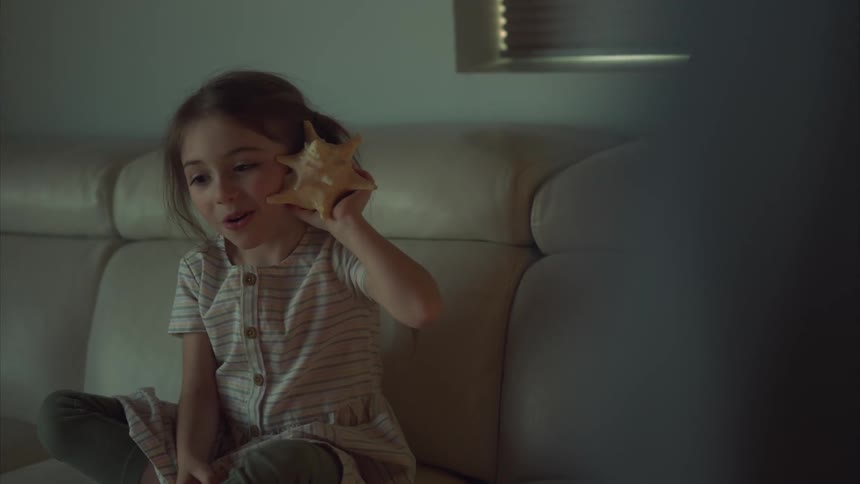 Little girl sitting on the couch and interacting with a seashell in her ear