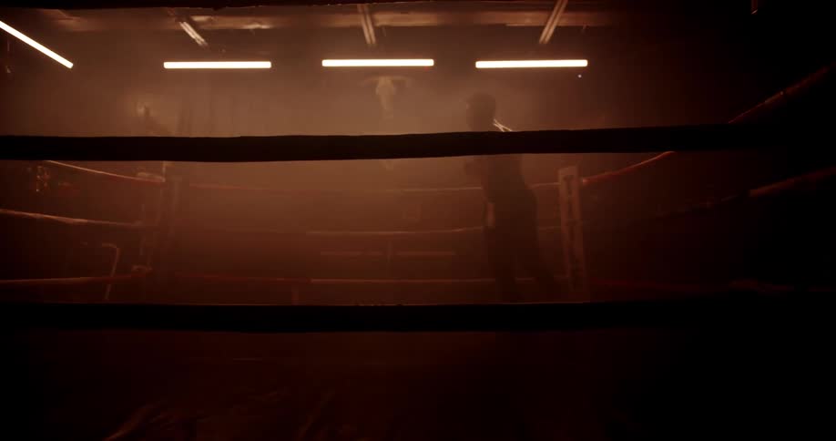 A shirtless man trains in boxing within a ring in a dimly lit gym