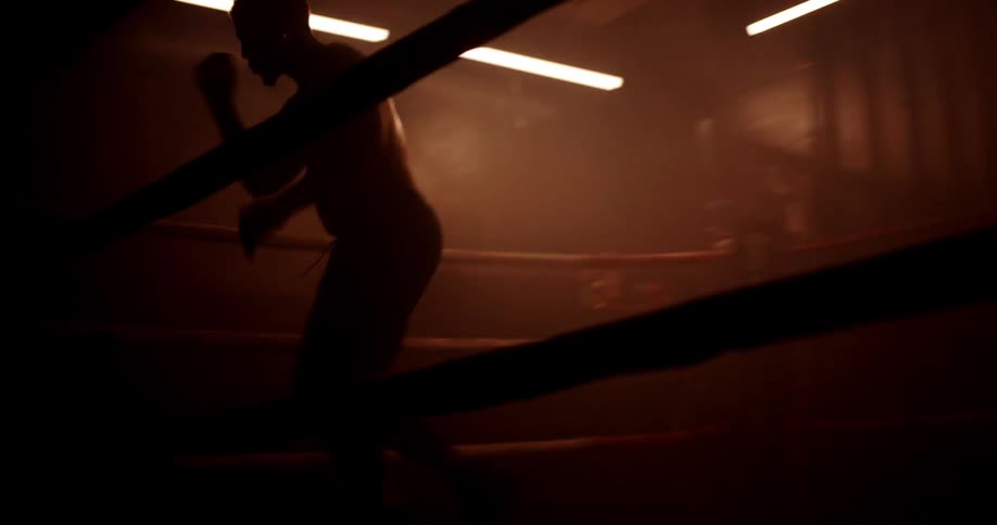 A boxer trains alone in a dimly lit gym practicing on the ring