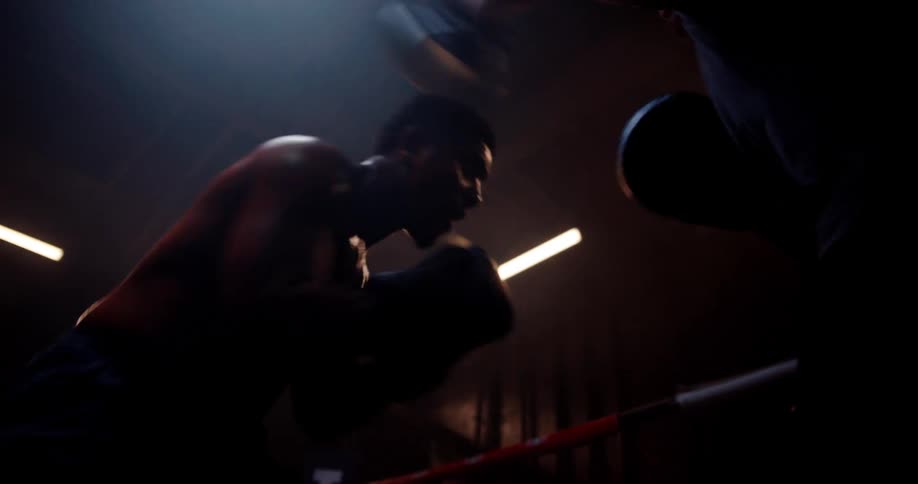 A boxer trains with a sparring coach in a dimly lit gym practicing on the ring