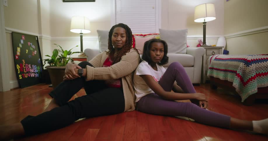 Mother and daughter sit back to back on a wooden floor in a cozy living room featuring two lit lamps a white couch a colorful blanket and a small plant