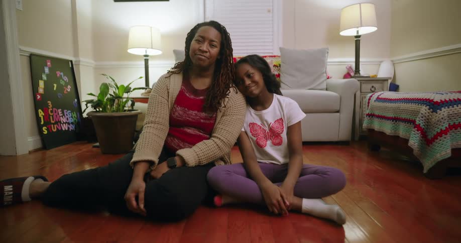 Mother and daughter sitting on the floor of a sitting area smile and lean against each other
