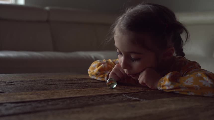 A girl sits on the floor of a living room gazing at a marble placed on a wooden table and pushing it away