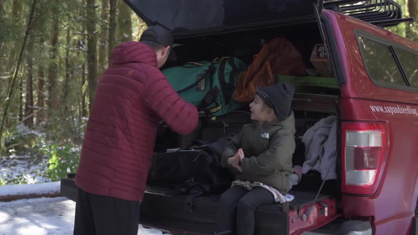 A little boy sitting in the trunk of a car and a man putting luggage in it