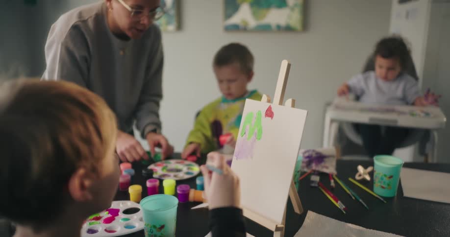 A group of children and an adult engaged in a painting activity with art supplies like brushes paint palettes and paper on a table