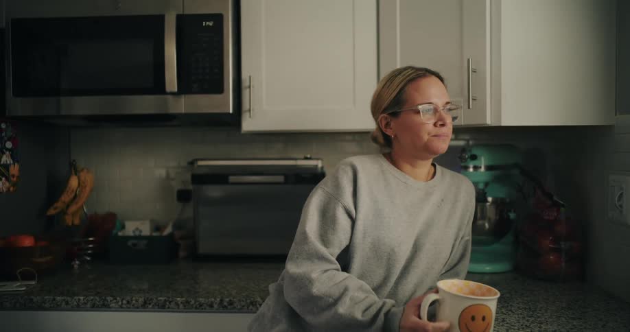 A woman is standing in her kitchen and drinking coffee