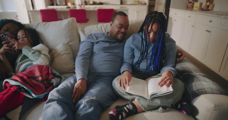 A family sits together on a large couch in the living room some using smartphones while others are reading