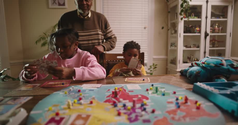 A grandfather watches as his two grandchildren play a board game at a table