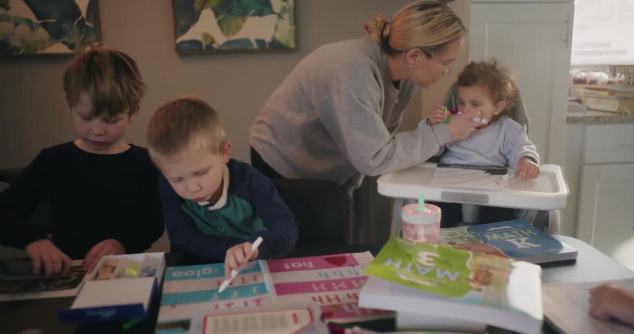 A mother tending to their children while they do homework over a cluttered table at home