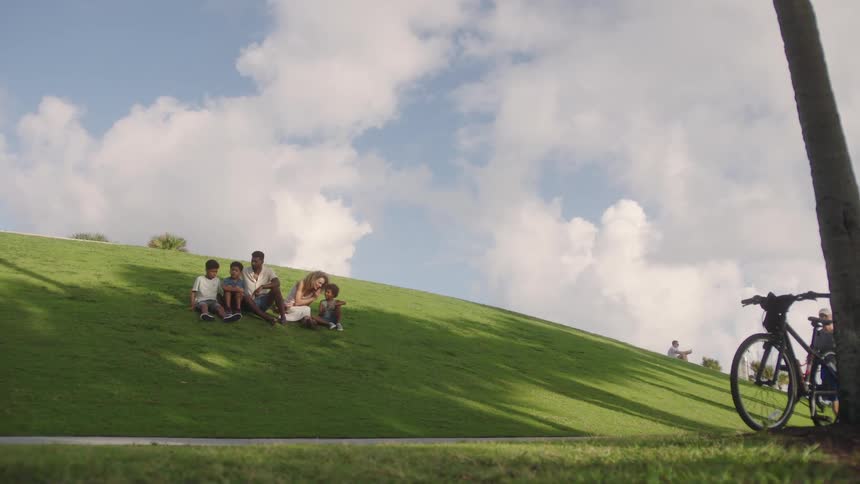 A family of five sitting on a lush green hill under a partly cloudy sky