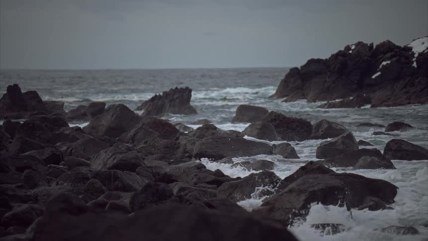 Waves crash vigorously against a rugged shoreline scattered with dark rocks where small patches of snow cling to the stony edges under a moody overcast sky looming overhead
