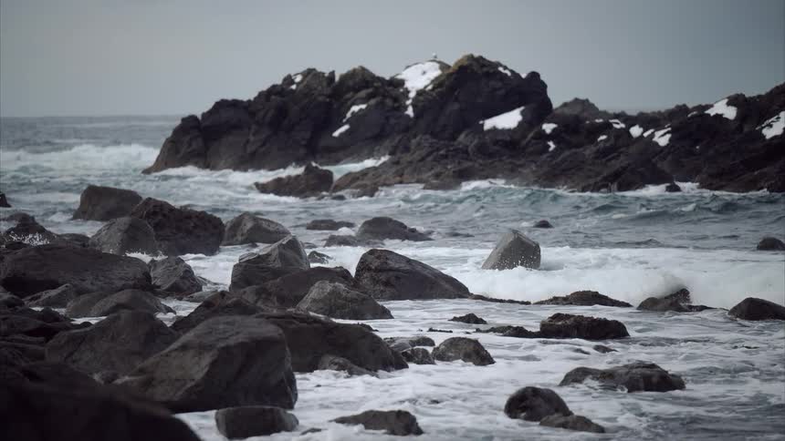 Waves crash against dark rocky outcroppings some dusted with snow in a turbulent sea under an overcast sky