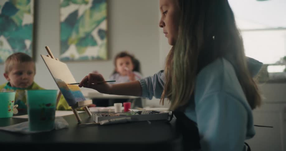 A girl paints on a small canvas set on an easel surrounded by art supplies and a green container on a table