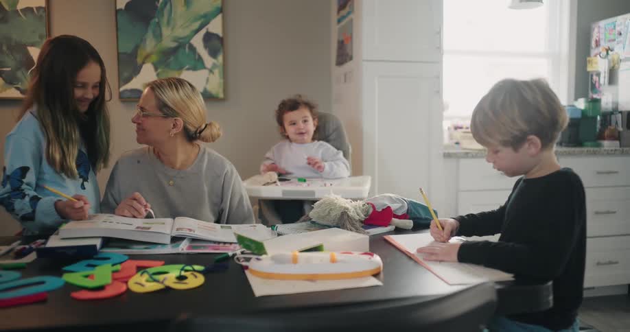 A busy mother helping her children with homework at a dining table at home on a sunny day