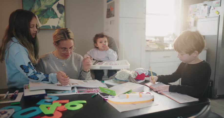 The mother is sitting at the kitchen table with one kids beside her in a high chair another girl standing beside her asking for help and her one son is sitting and working on a paper and the other son is playing with numbers